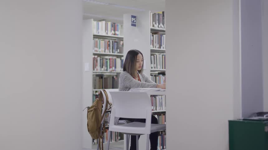 A young asian woman sitting at a table in a library takes notes while flipping through a large book