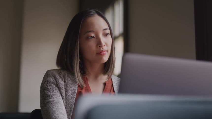 Asian teenager girl with straight hair and a grey long sleeve coat sitting at a table uses a laptop