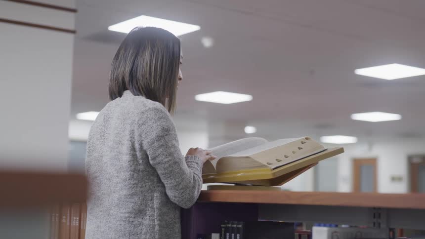 A young asian woman flips through the pages of an encyclopedia on top of a bookshelf in a library