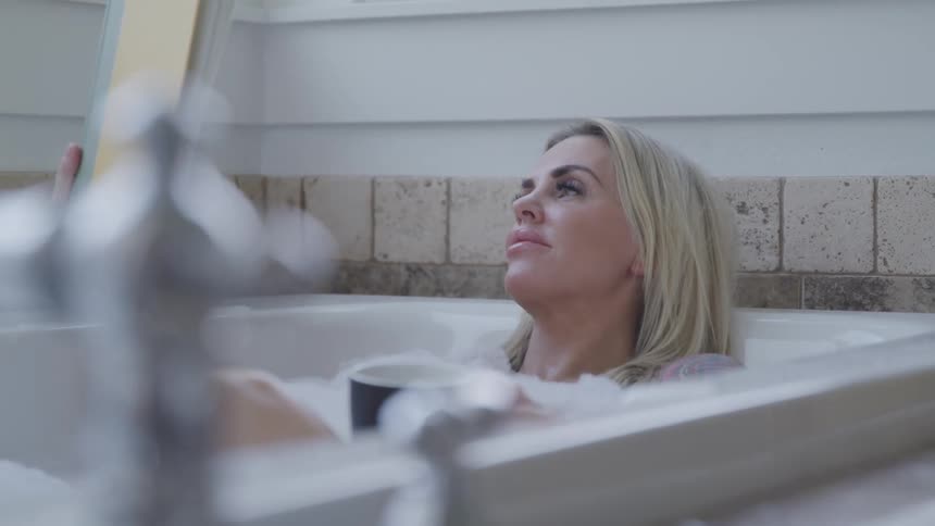 A woman sits in a bubble bath holding a coffee mug and a book