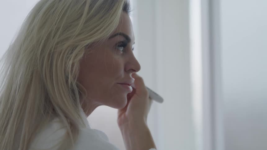 A woman in a bathrobe uses a makeup brush to apply make up