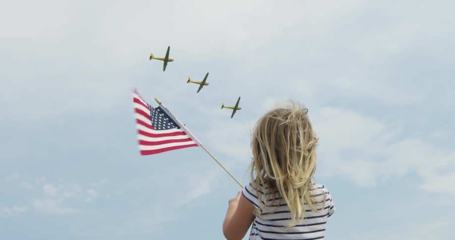 A girl holds up an american flag while planes fly in the sky