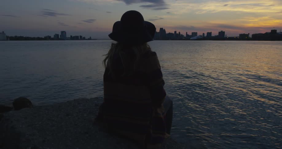 A woman sitting next to a body of water in an urban area at sunset
