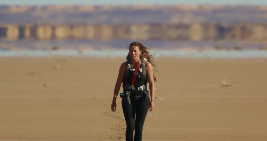 Woman walks across sand carrying camera on strap as air shimmers over cliffs on horizon