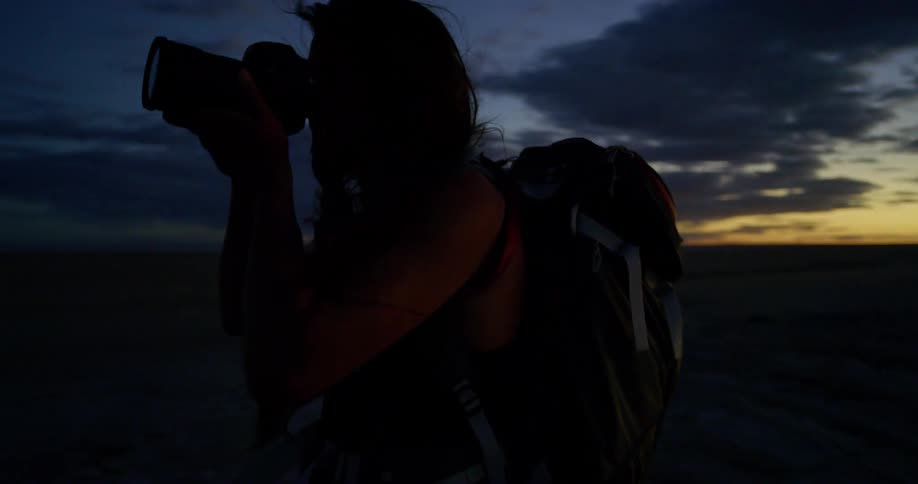 A female photographer walks away from the rising sun and raises her camera to photograph the darkness beyond