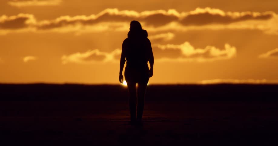 A silhouette of a person walking in front of a sliver of setting sun