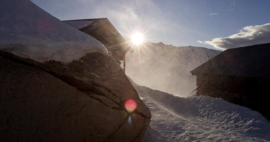 Wind blowing snow around a pair of wooden houses in the mountains on a sunny day
