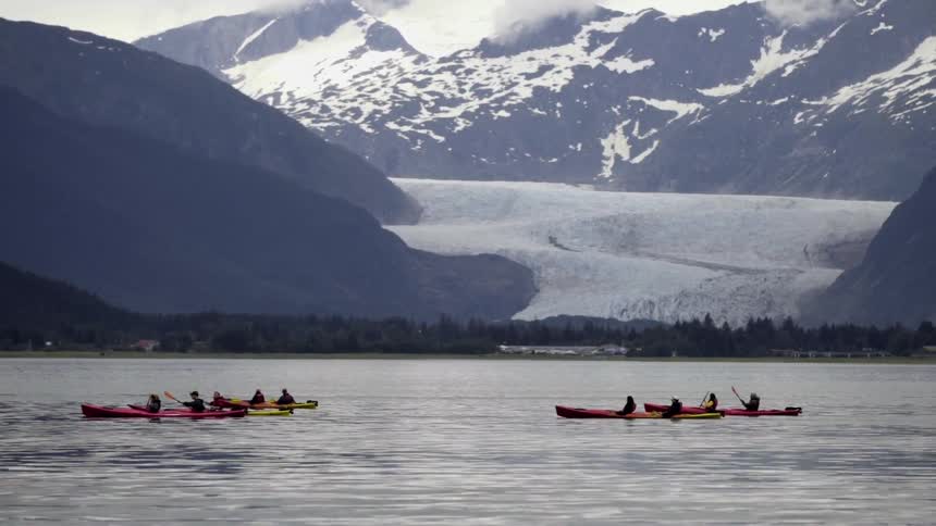 Several multi-person kayaks moving on a body of water with snow-covered mountains in the background