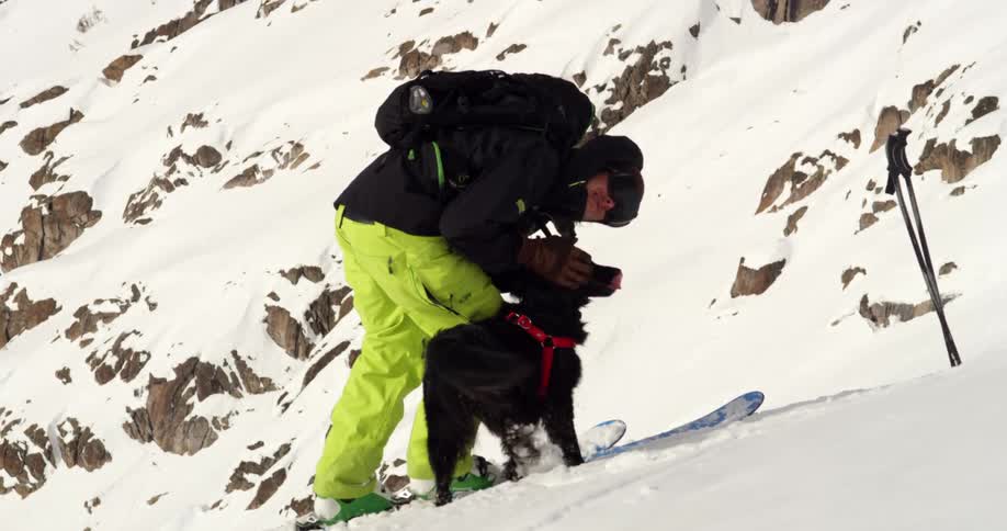 Man wearing winter and ski gear cuddles with his black pet dog on the snow in the mountains