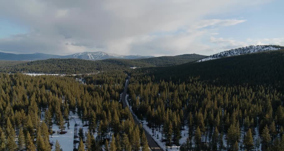 A road surrounded by trees and snow with a mountain range the sky and clouds in the background