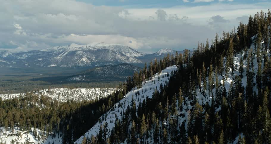 A hill with trees covered with snow with a mountain range and the sky in the background
