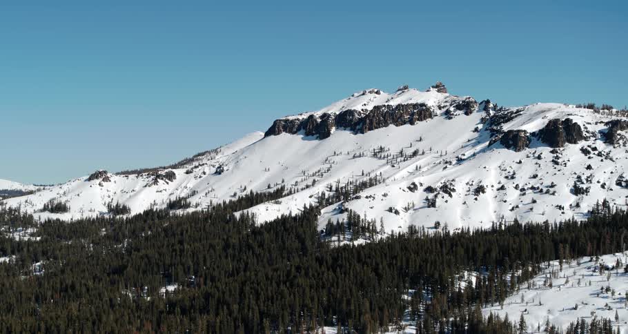 Snow covers the slope of a mountain range and the ground below a forest of dense trees
