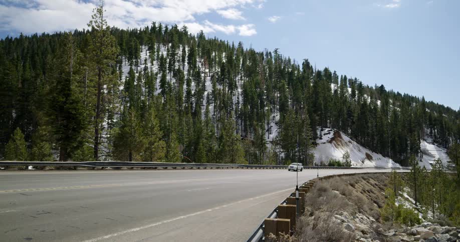 A vehicle travels down a road along a mountain covered in snow and evergreen trees