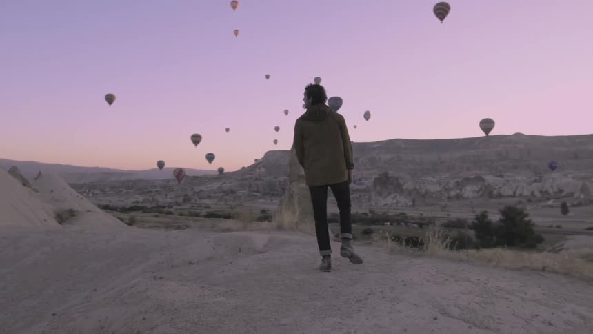 A man climbs on a hill and looks around at the hot air balloons all over in the air