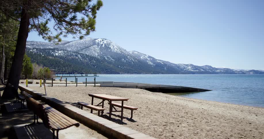 A table and bench next to the shore and trees with a mountain range and sky in the background