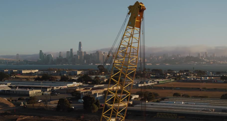 A construction crane in front of buildings with cars driving past and a city skyline in the distance