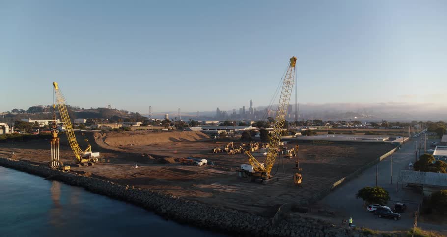 Cranes stand on bare lot near water bordering hill with fog covered city in background