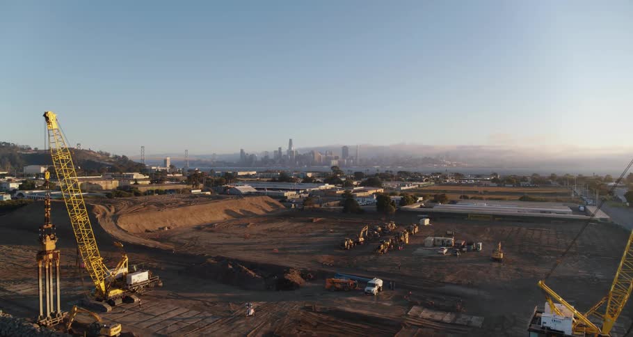Cranes and construction equipment sit on a large dirt oil field next to the ocean