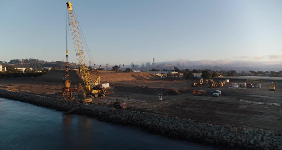 Construction cranes near a shore with trees the sky and clouds in the background
