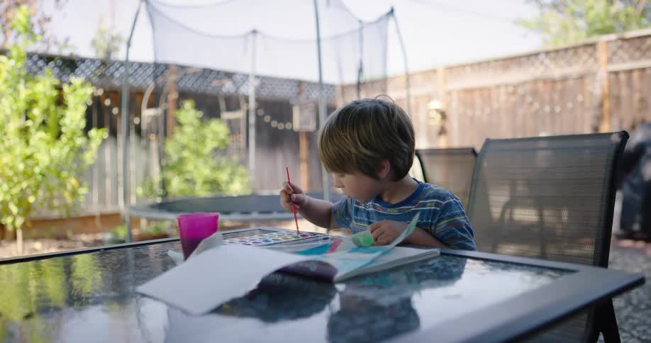 A little boy sitting at a table in a backyard paints in a pad of paper