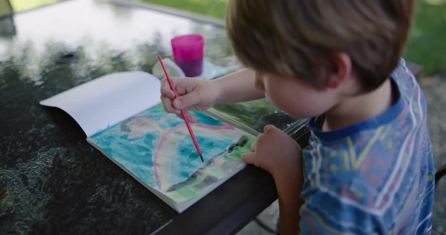 A little boy sitting at a table in a back yard paints a picture