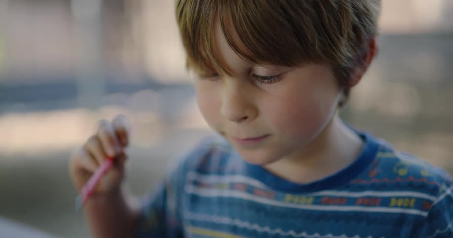 A boy sits at a table and looks at his painting as he holds a small paintbrush
