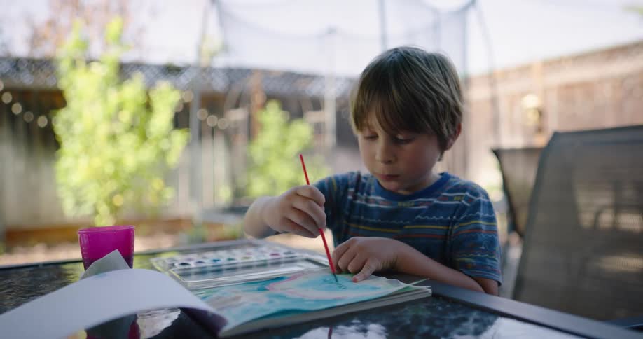 A young boy sits outside at a patio table and uses water color on a book