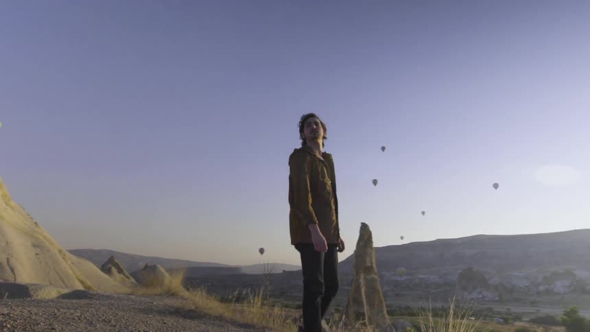 Man standing in a desolate area looking all around at the many hot air balloons floating in the sky at sunrise