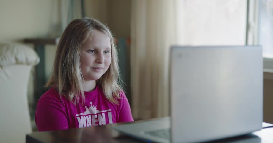 Female child sitting at the table in front of the screen of a laptop smiles and waves as she video chats