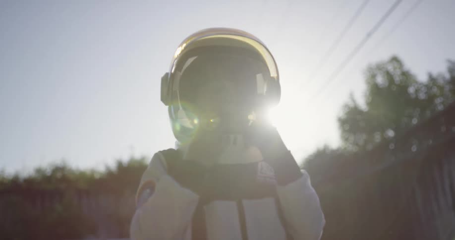 A boy in an astronaut costume stands outdoors within a fenced off yard during the day