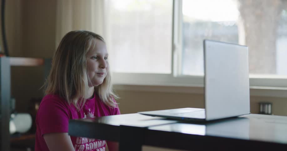 Little girl with blonde hair and a pink t shirt uses a laptop and communicates with a person over the internet using a video call