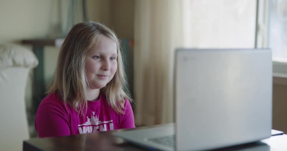 A girl sits at a table in front of a laptop appearing to have a video call