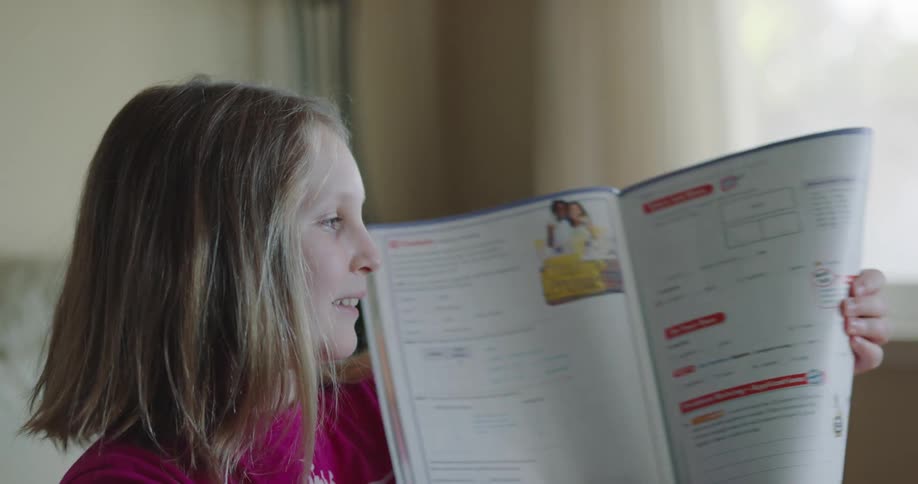 A girl sitting in her living room talks and lifts her math workbook to show it to someone out of frame