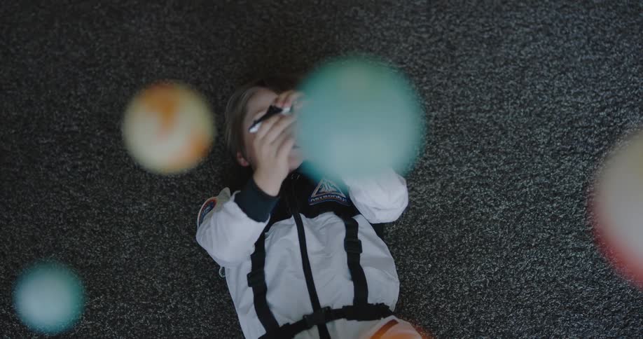 A boy in an astronaut costume lies on his back on the carpet as he plays with a space shuttle toy