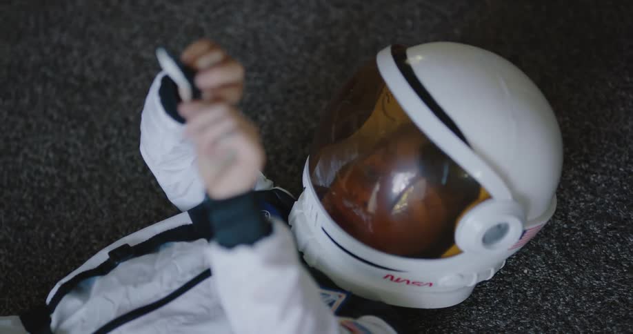 A little boy in a space suit lying on the floor at home plays with a toy spaceship