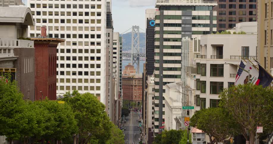 Car drives down sloping street through downtown toward suspension bridge and mountain