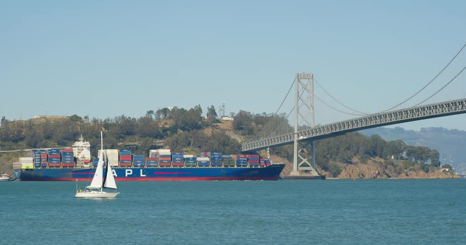 A large freighter and a small sailboat travel under a bridge on the ocean on a sunny day