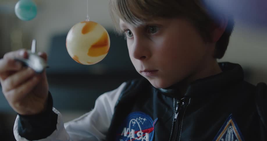 A little boy plays inside with a toy space ship as toy planets hang down from the ceiling