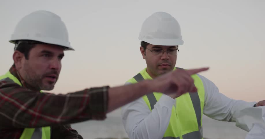 A pair of constructions walks review and discuss a blueprint while standing outdoors in safety gear during the day