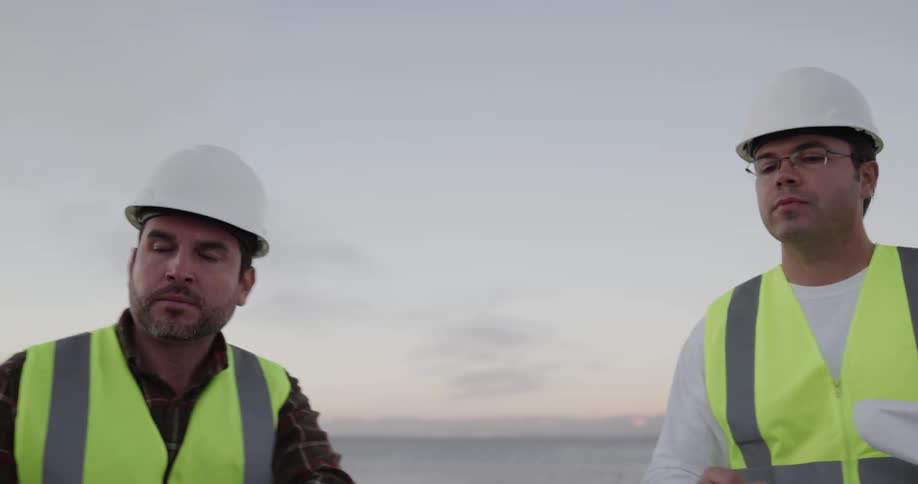 Two engineers wearing white hard hats discuss over blueprints outdoors at sunset