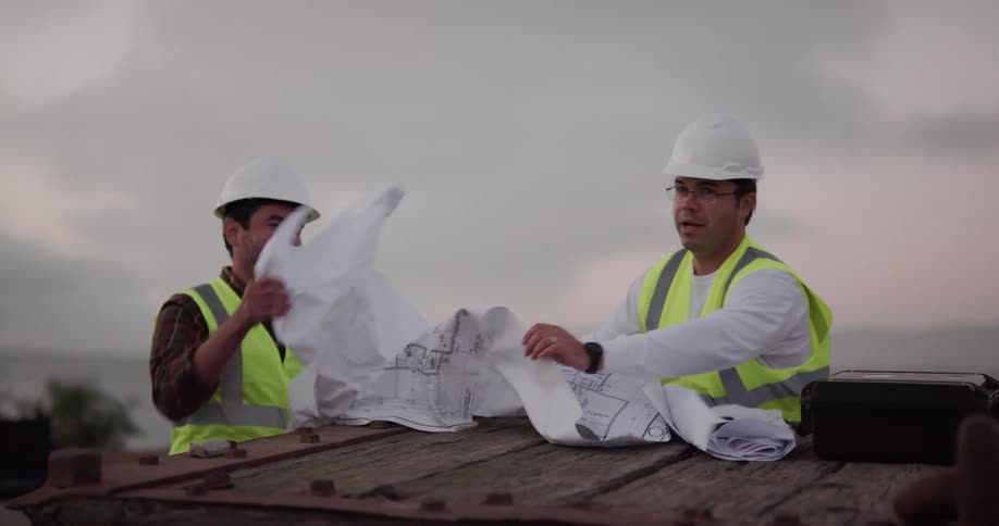 Two construction workers talk and try to gather up blueprints as the wind blows them