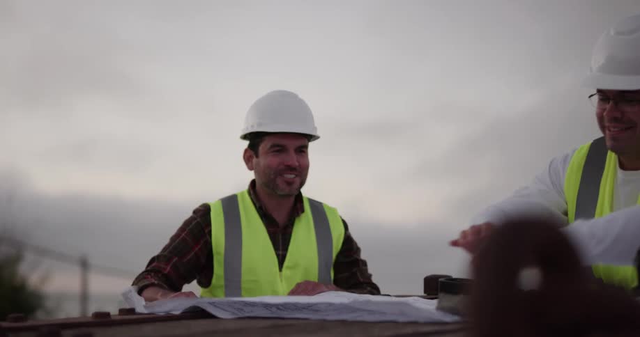 Two man in bright yellow vests and white hard hats talks while reviewing construction plans
