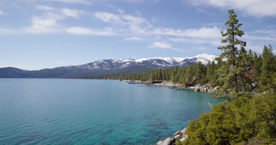 A large and serene lake is surrounded by a dense forest nearby to snow capped mountains