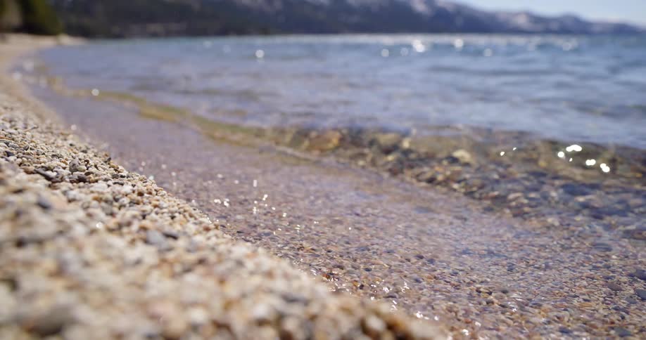 Clear ocean waves wash onto a gravelly shore of a beach on a sunny day