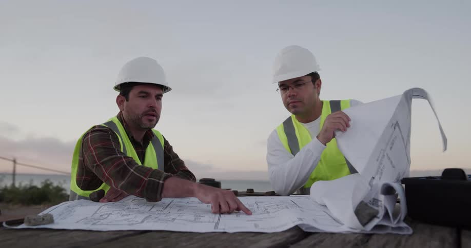 Two construction workers with hard hats and vests talking and looking over blueprints with the sky and clouds in the background