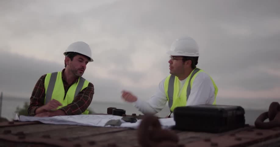 A pair of construction workers standing outdoors on a windy day review and discuss a blueprint as it shakes in the window