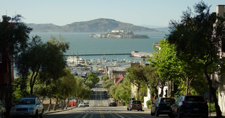 A street lined with parked vehicles leading down a hill in a city on the coast on a sunny day