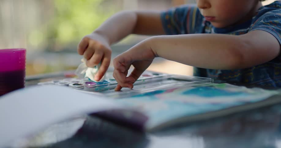 A boy cleans his watercolor paint set with a napkin as he sits at a table