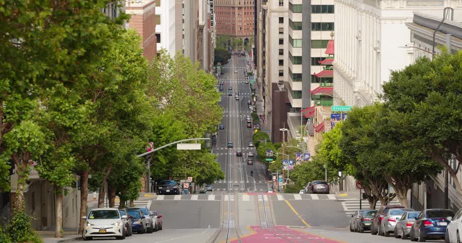 A wide street lined with parked cars and skyscrapers on a hill in a metropolis during the day