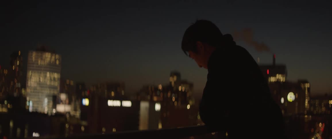 A man leans on the railing of a balcony overlooking a city at night